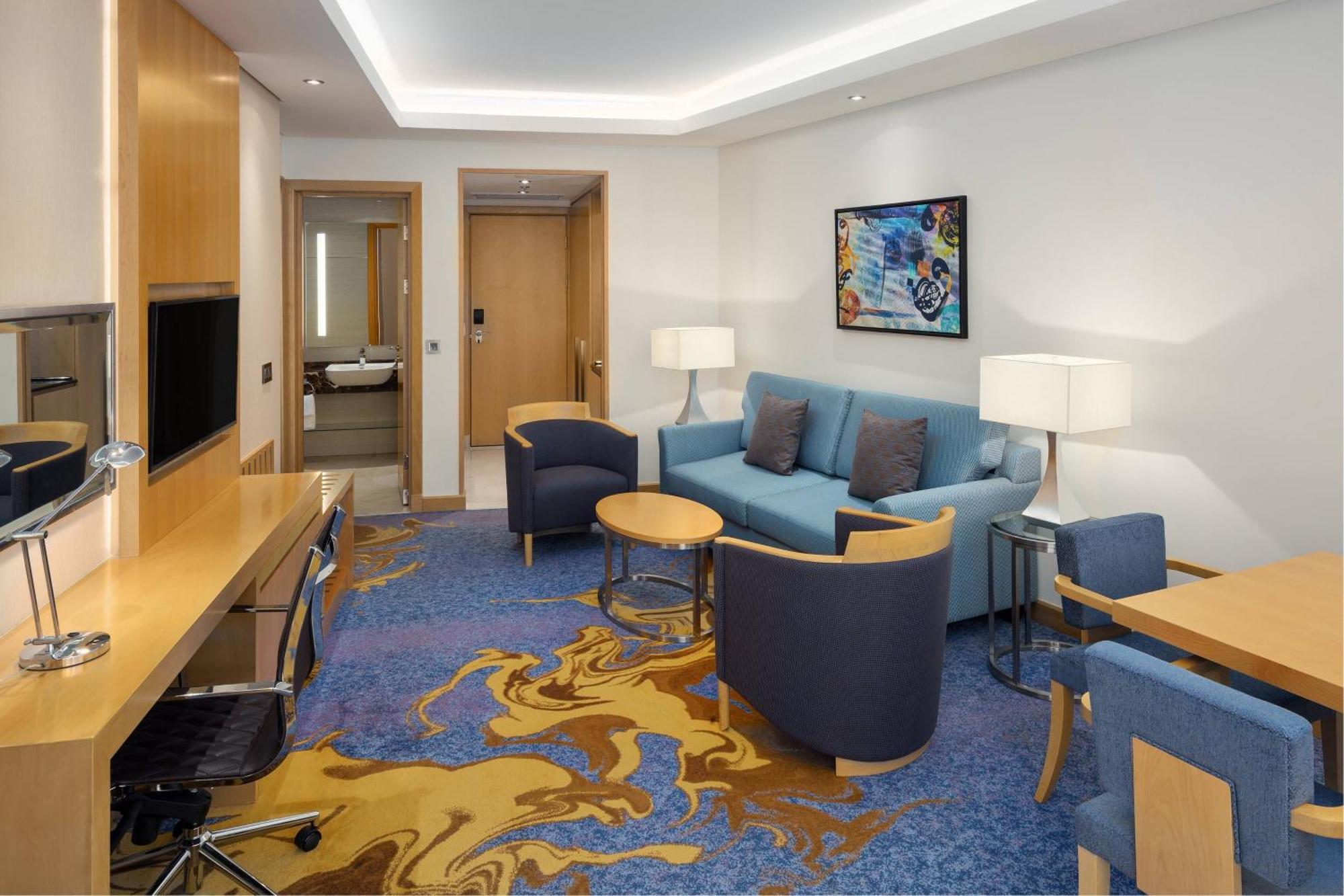쉐라톤 마카 자발 알 카바 호텔 메카 외부 사진 The photo displays a modern hotel room or suite. The room features a comfortable seating area with a blue couch and two blue armchairs surrounding a small round table. A wooden desk with a black office chair is positioned against one wall, accompanie