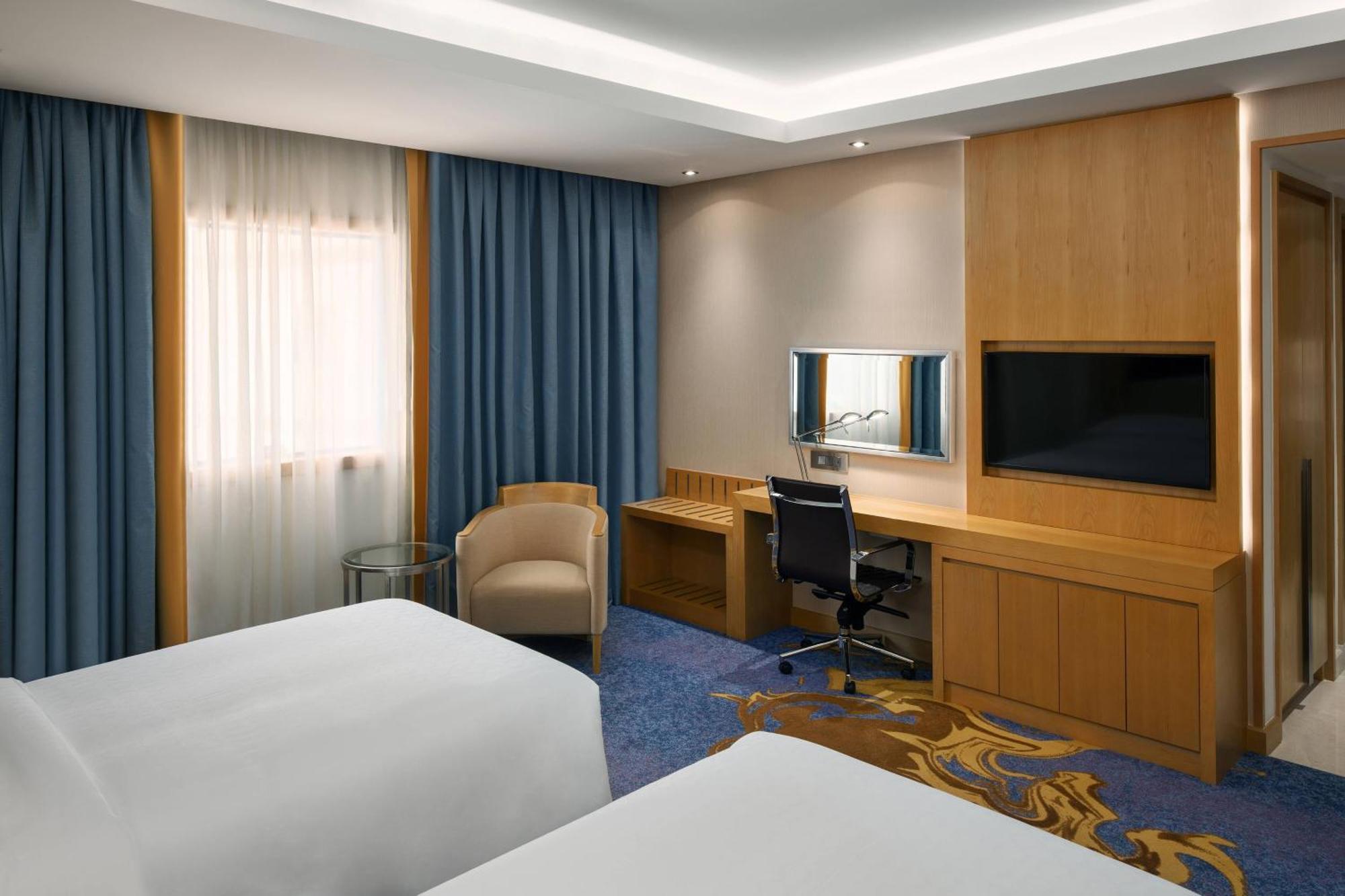 쉐라톤 마카 자발 알 카바 호텔 메카 외부 사진 The photo shows a hotel room featuring two beds with white linens. There are large windows covered with blue curtains that allow natural light to enter. On one side of the room, there is a wooden workstation with a chair and a mirror, along with a fl