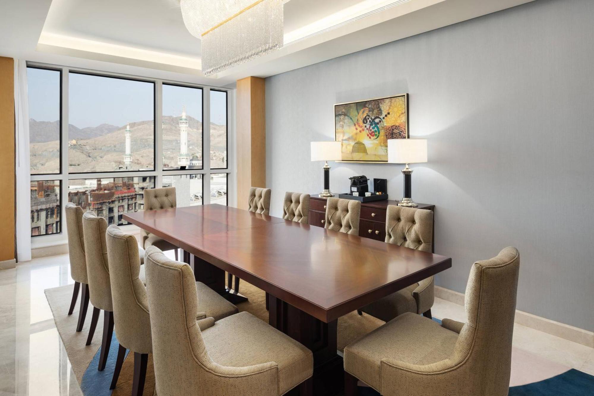 쉐라톤 마카 자발 알 카바 호텔 메카 외부 사진 The photo shows a modern conference room with a large wooden table surrounded by upholstered chairs. The room is well-lit, featuring large windows that offer a view of the outdoors, possibly with mountains in the background. On one wall, there is a p